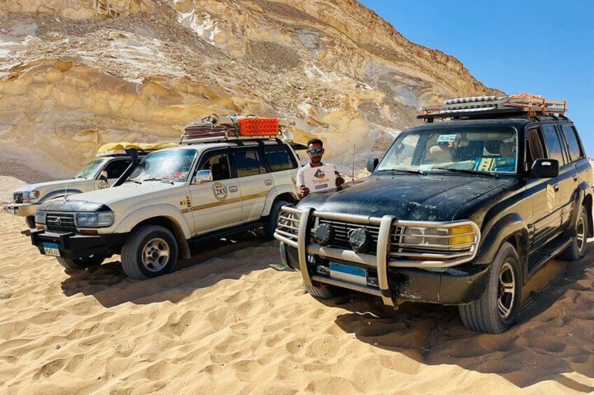 Two off-road vehicles parked in a desert with rocky terrain in the background. 3