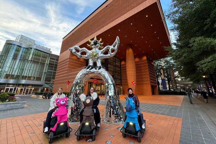 Tres personas en patinetes, una escultura de árbol grande y un edificio con techo rojo. 3