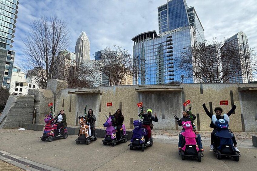 Un grupo de personas en scooters eléctricos alineadas frente a un edificio de fachada de piedra y banderas rojas. 1