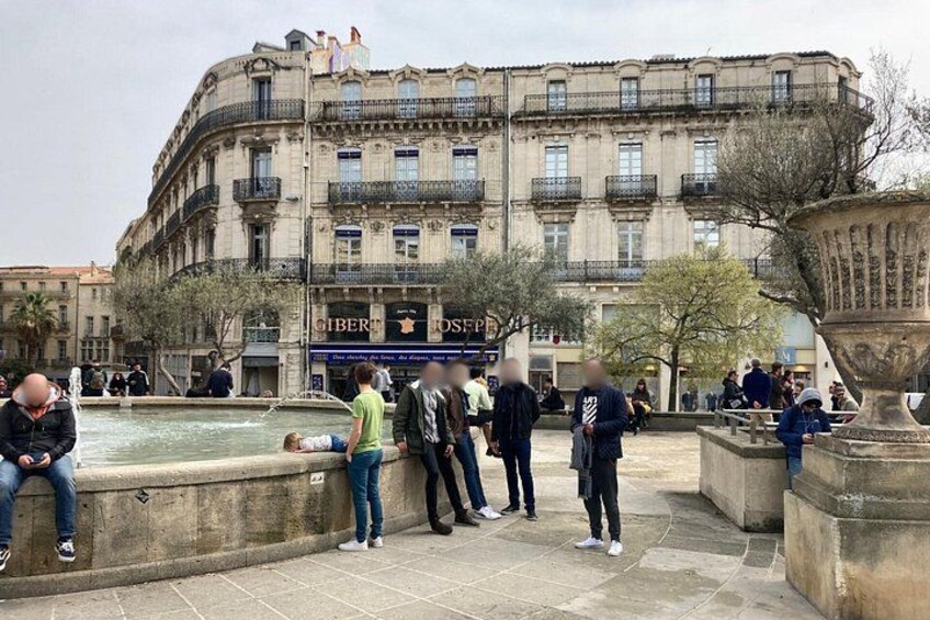 Une place publique avec une fontaine, des gens assis et debout, et un bâtiment historique doté de balcons. 5