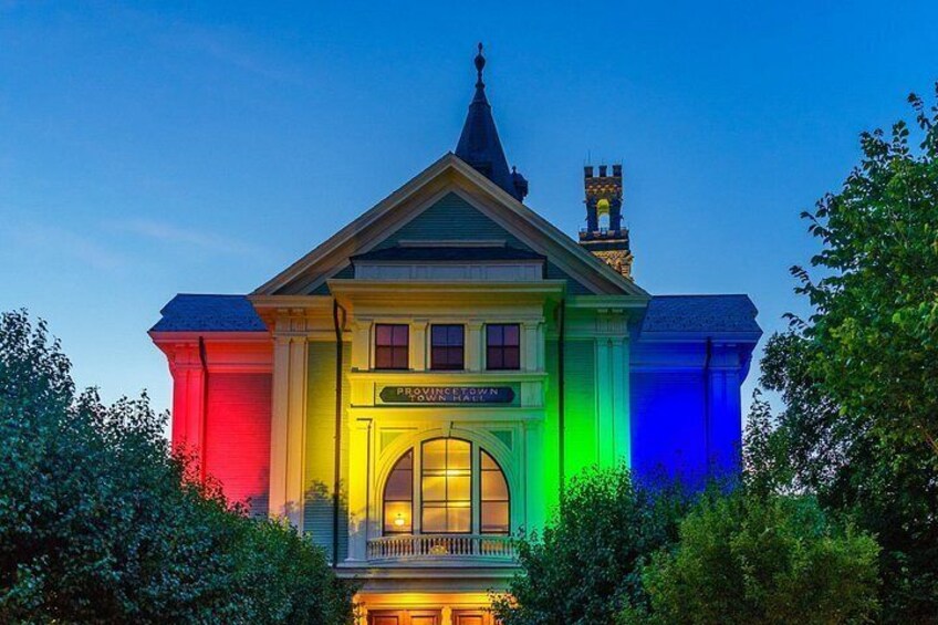 A building with a pointed roof and a clock tower, illuminated with multicolored lights. 2