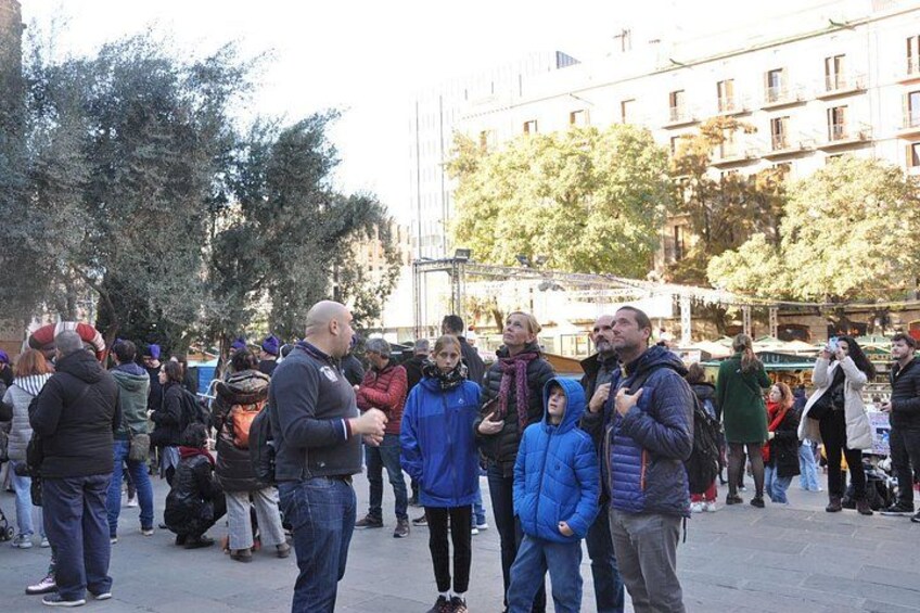 A group of people standing outdoors in a public square, with buildings and trees in the background. 2