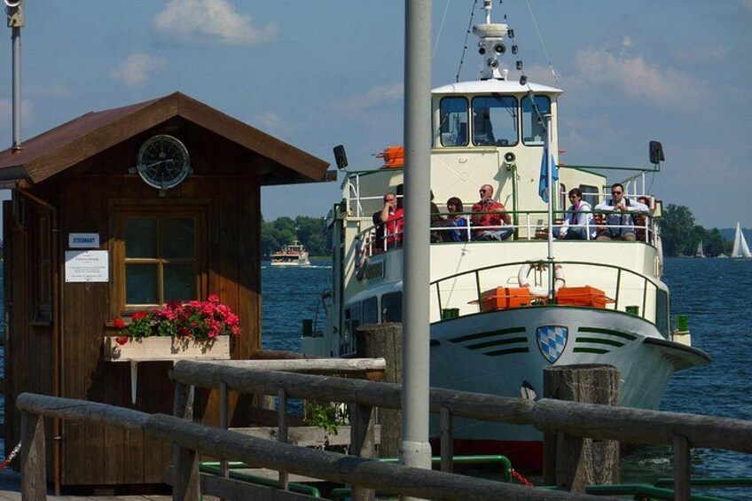 A wooden building with a clock, a boat with passengers, and a sailboat in the background. 5