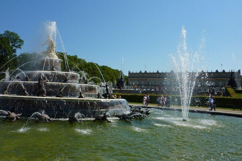 A multi-tiered fountain with water jets, surrounded by a well-maintained garden and a large building in the background. 3
