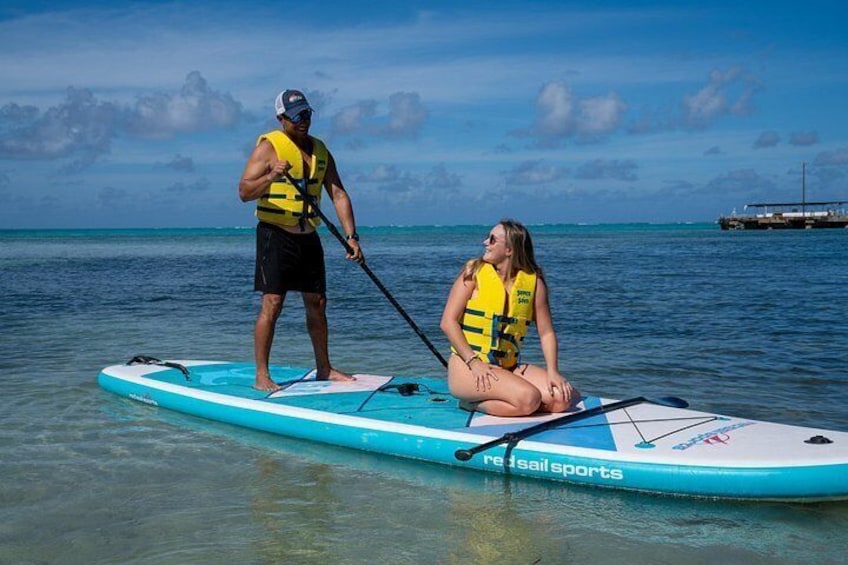 Deux personnes pratiquent le stand-up paddle sur une planche turquoise, avec un ciel bleu clair et une mer calme en arrière-plan. 3