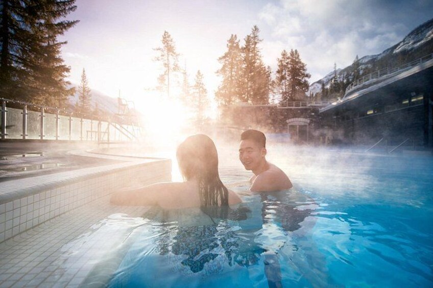 A hot spring with two people in the water, surrounded by trees and a building in the background. 1