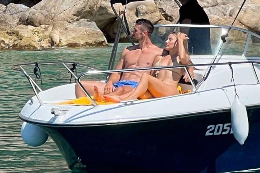 A boat with two people, one in a blue swimsuit and the other in a yellow swimsuit, on calm water with rocks in the background. 1