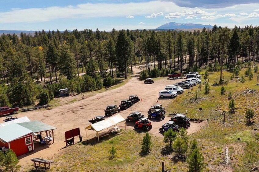 A parking lot with vehicles, a red building, and a forest in the background. 3