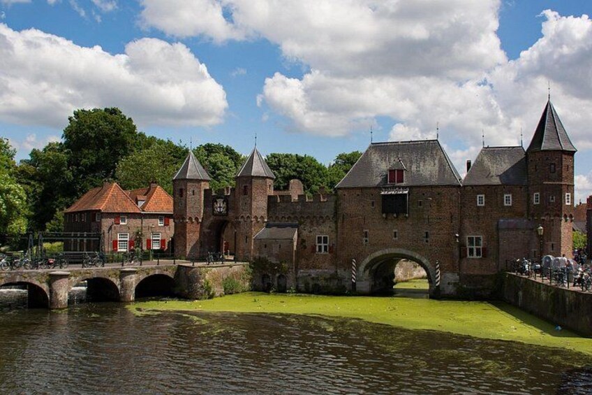 Een historisch bakstenen gebouw met een brug, een kanaal en groen op de achtergrond. 1