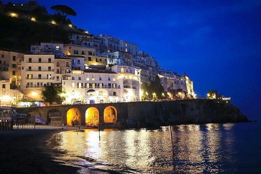 A coastal town at night with illuminated buildings and a bridge over the water. 1