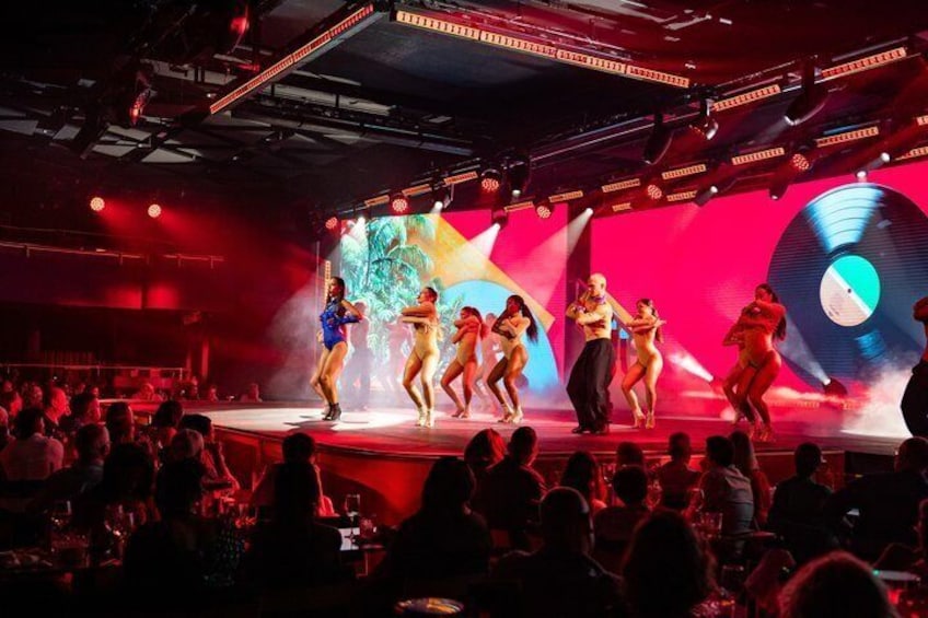 Una función en escenario con bailarines vestidos con trajes coloridos, un gráfico de un disco de vinilo grande y un público en un recinto con poca luz. 3