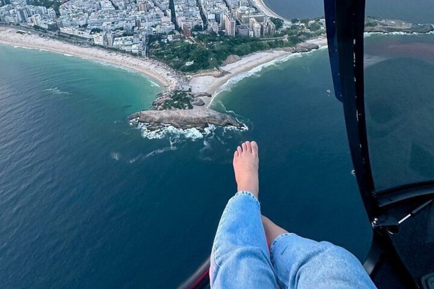A person's feet dangling over a cliff, overlooking a coastal city with buildings and a beach. 5