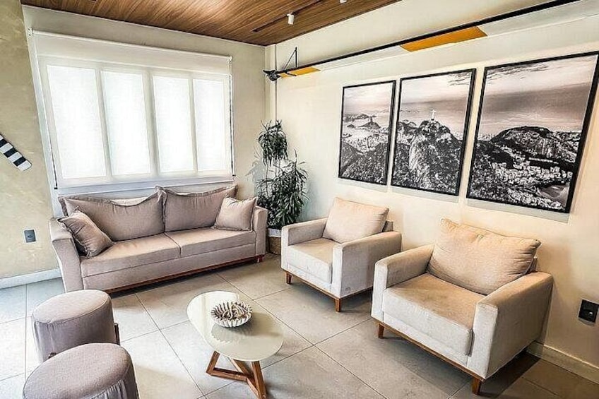 A living room with a beige sofa, two armchairs, a coffee table, and a large window with blinds. There are three framed photographs on the wall. 2