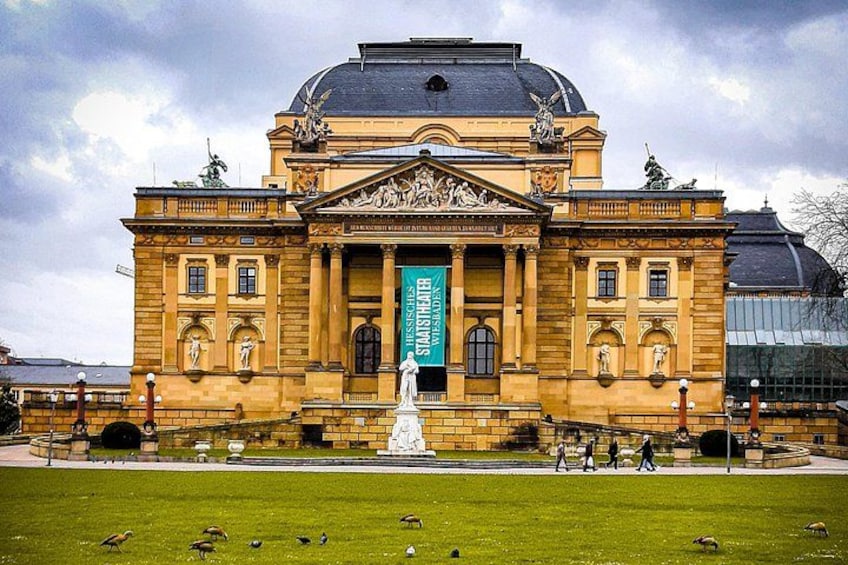 A historic building with a central statue, surrounded by a grassy area and a few people walking. 2