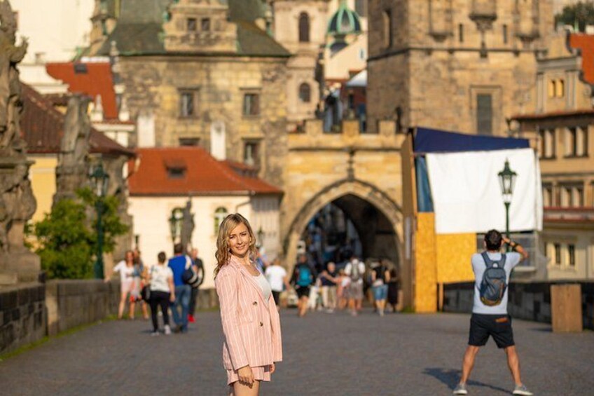 A person standing in front of a historic stone bridge with arches, a flag, and a person taking a photo in the background. 3