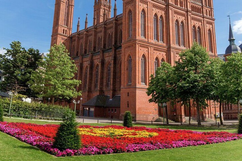 A large red brick church with a tall tower and a colorful flower bed in the foreground. 4