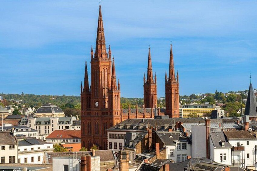 A cityscape with prominent red brick Gothic-style towers, surrounded by modern buildings and a clear blue sky. 1