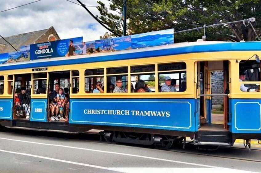 Eine blaue und gelbe Straßenbahn mit der Aufschrift 'Christchurch Tramways', Fahrgäste im Inneren und einem Schild mit der Aufschrift 'Gondola' darüber. 3