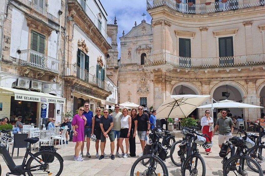 A group of people standing in a public square with bicycles parked around them, and historic buildings in the background. 1