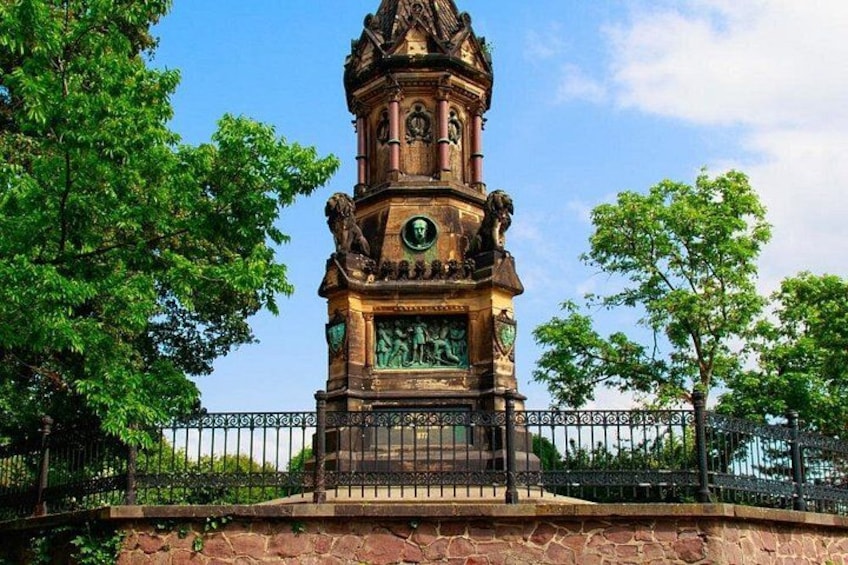 A clock tower with intricate carvings and a statue, surrounded by trees and a fence. 4
