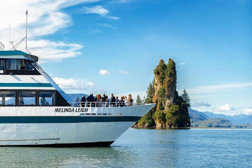 Een boot genaamd MELINDA LEIGH met passagiers, een rotsachtig eiland met bomen en een bergachtig landschap op de achtergrond. 1
