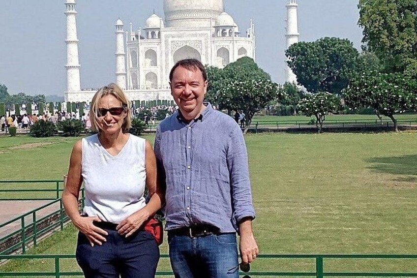 Dos personas de pie frente al Taj Mahal, con un césped verde y árboles al fondo. 3