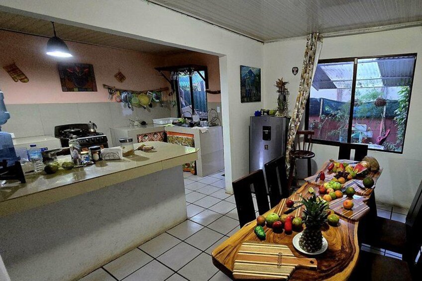 A kitchen with a dining table set with fruits, a refrigerator, and a window with a view of the outdoors. 2