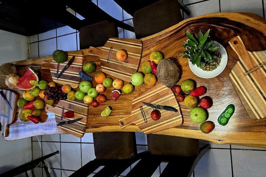 A wooden cutting board with various fruits and vegetables, a knife, and a bowl of pineapple on a tiled floor. 1