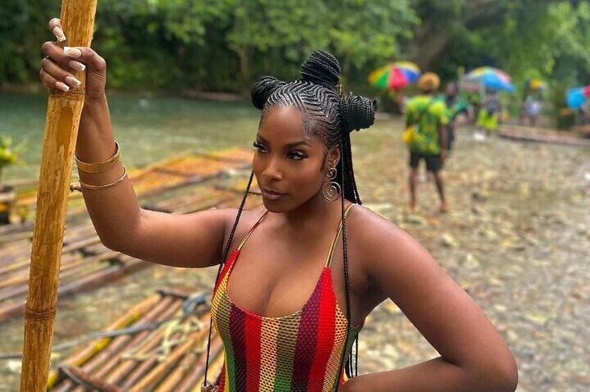 A person in a colorful swimsuit holding a bamboo pole, standing on a wooden platform by a river with people in the background. 2