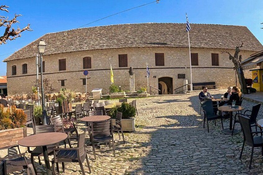 Een gebouw van steen met een pannendak, een terras met tafels en stoelen, en een vlaggenmast met vlaggen. 5