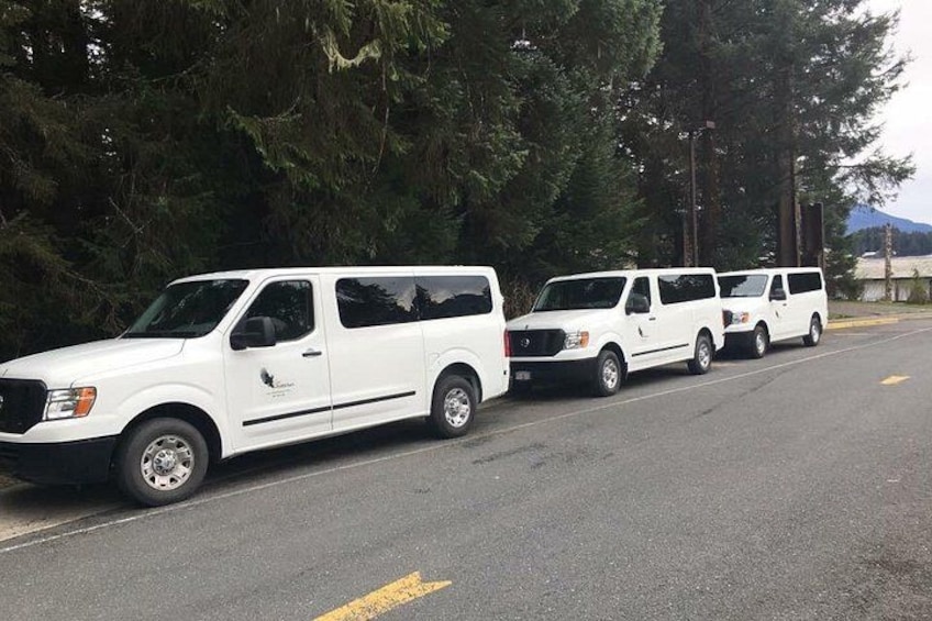 En rad vita skåpbilar står parkerade längs en väg, med träd och berg i bakgrunden. 3
