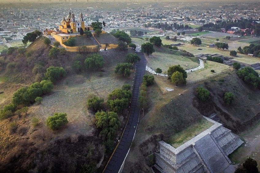 An aerial view of a hilltop fortress with a road leading up to it, surrounded by greenery and a distant cityscape. 3