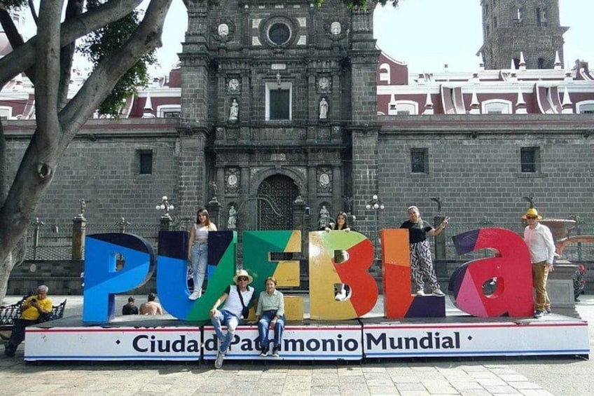 A group of people standing in front of a large sign with the word 'Puebla' and a historic building in the background. 2