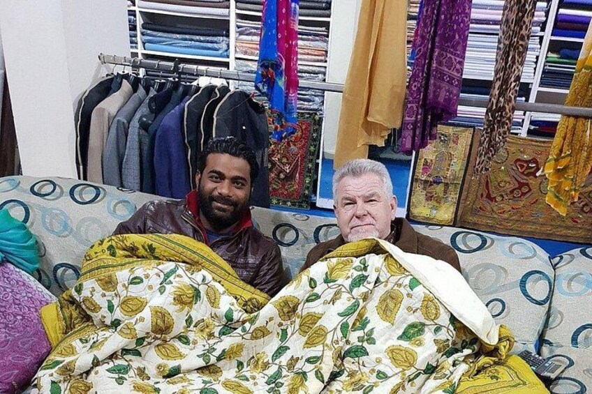 Dos personas sentadas en un sofá estampado con una ventana y perchas de ropa al fondo. 1