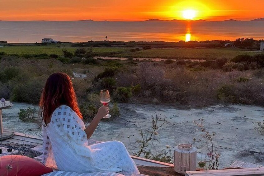 A person sitting on a chair holding a glass, overlooking a sunset over a coastal landscape. 1