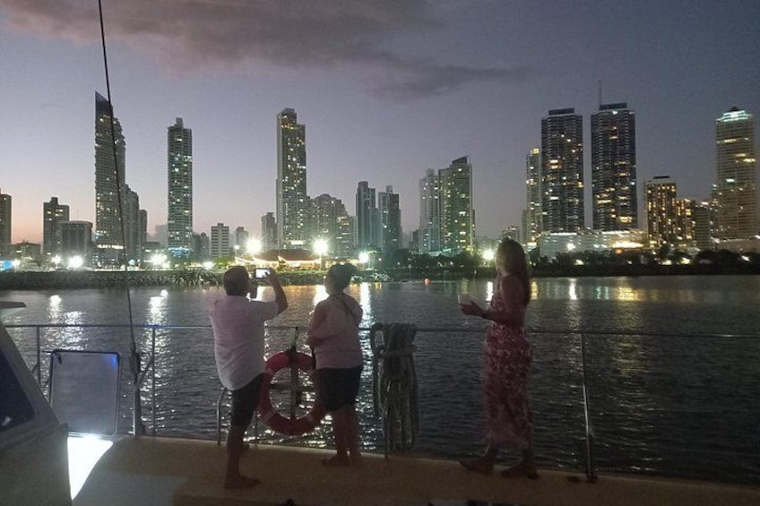 A city skyline at dusk with illuminated buildings, a body of water, and people on a boat. 4