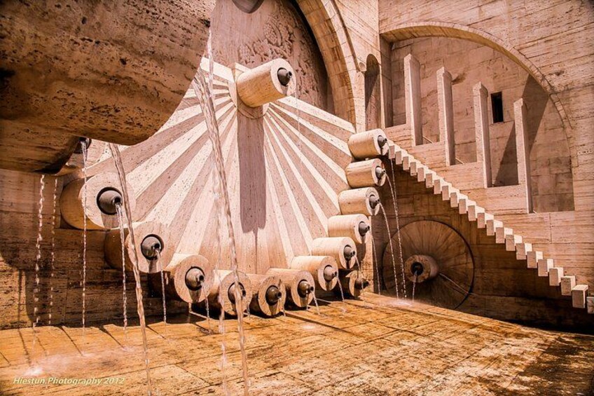 A large, ornate water wheel with a staircase leading up to it, set against a stone wall. 3