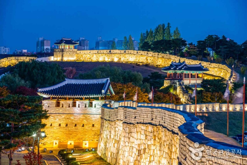 A historic stone wall with traditional Korean architecture, including a pavilion and a curved structure, illuminated at dusk. 2