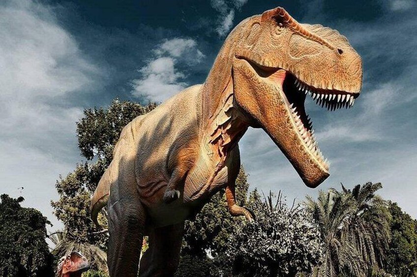 A life-sized model of a Tyrannosaurus rex dinosaur in a natural setting with trees and a clear sky. 4