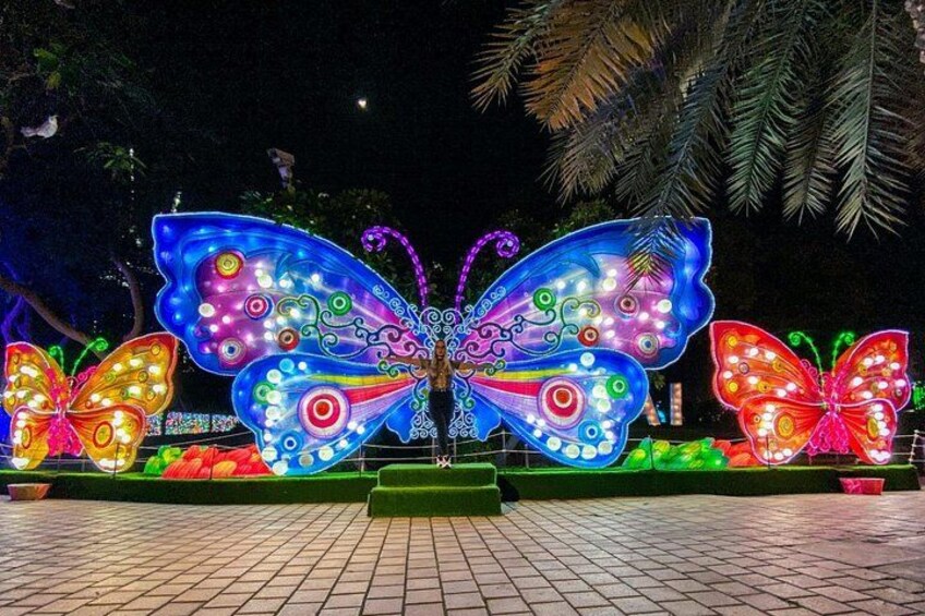 A large illuminated butterfly sculpture with intricate patterns and lights, set against a backdrop of palm trees and a paved area. 3