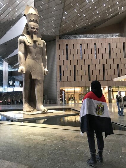Une grande statue d’un pharaon, dans un bâtiment moderne doté d’un mur orné de motifs, et une personne tenant un drapeau. 5