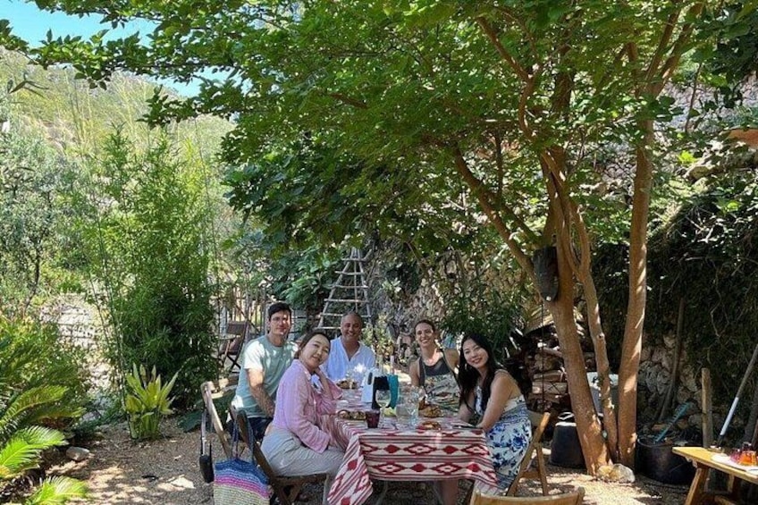 A group of people gathered around a table under a tree in a garden with plants and a wooden structure in the background. 4