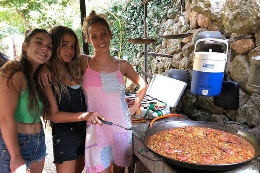 Three people near a large cooking pan, a Coleman water cooler, and a stone wall. 2