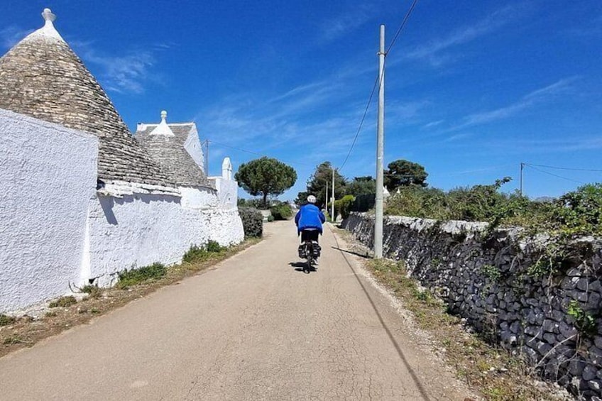 Une rue étroite bordée de bâtiments blancs et de murs de pierre, où l’on aperçoit un cycliste vêtu de bleu s’éloignant de la caméra. 3