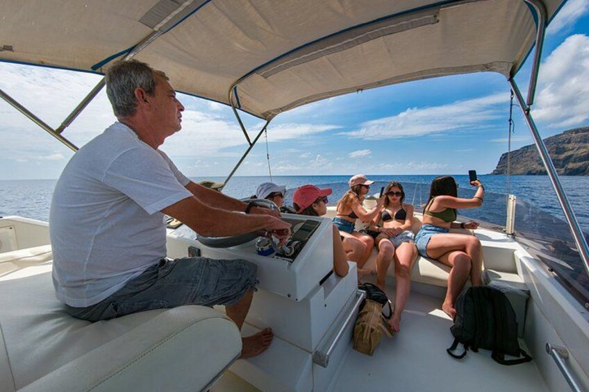 A boat with a man steering, passengers taking a selfie, and a clear view of the sea and distant cliffs. 5