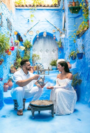 A couple seated in a blue-painted courtyard with a decorative wheel and traditional pottery. 3