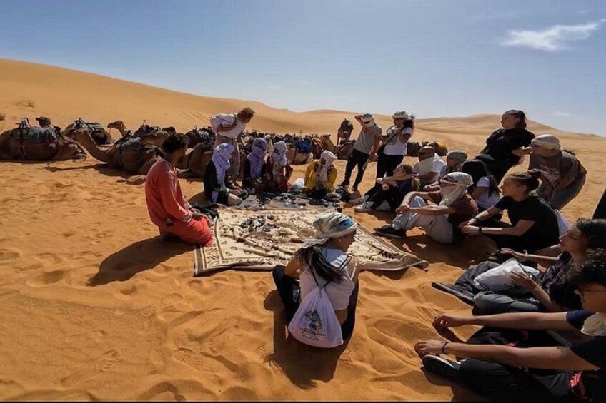 A group of people sitting and standing on a sandy area with camels in the background. 2