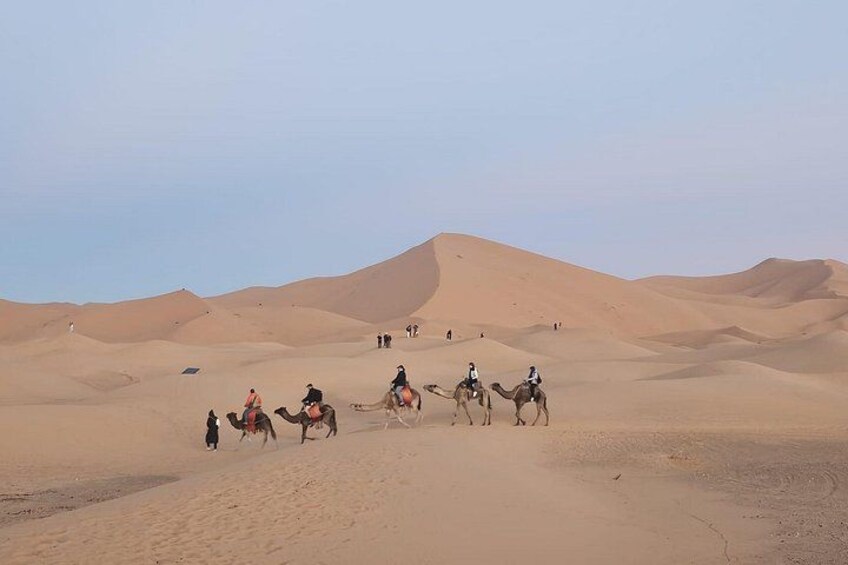 A group of people riding camels across a desert landscape with sand dunes in the background. 1