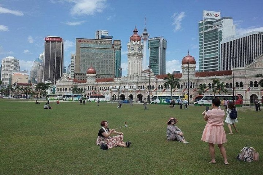 A grassy park with people sitting and standing, a large building with domes, and modern skyscrapers in the background. 5