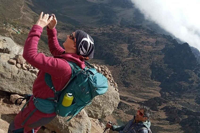 A person taking a photograph with a camera, wearing a backpack and a hat, standing on a rocky terrain with a mountainous background. 1
